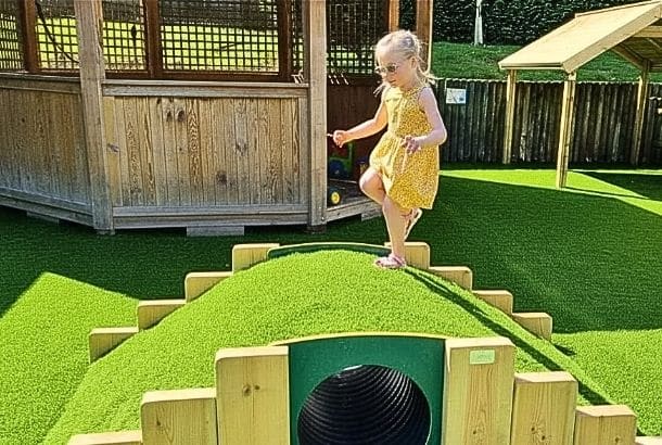 Girl in yellow dress running over a mound tunnel in a school playground covered in artificial grass