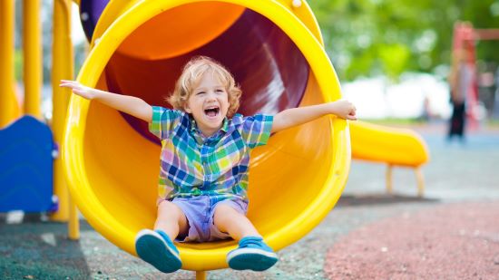 Young child in garish shirt popping out of a yellow tube slide with his arms out in celebration