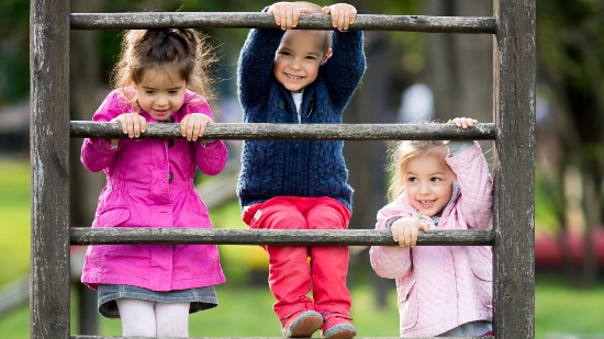 Three children climbing on outdoor equipment in the park, two of them smiling to the camera