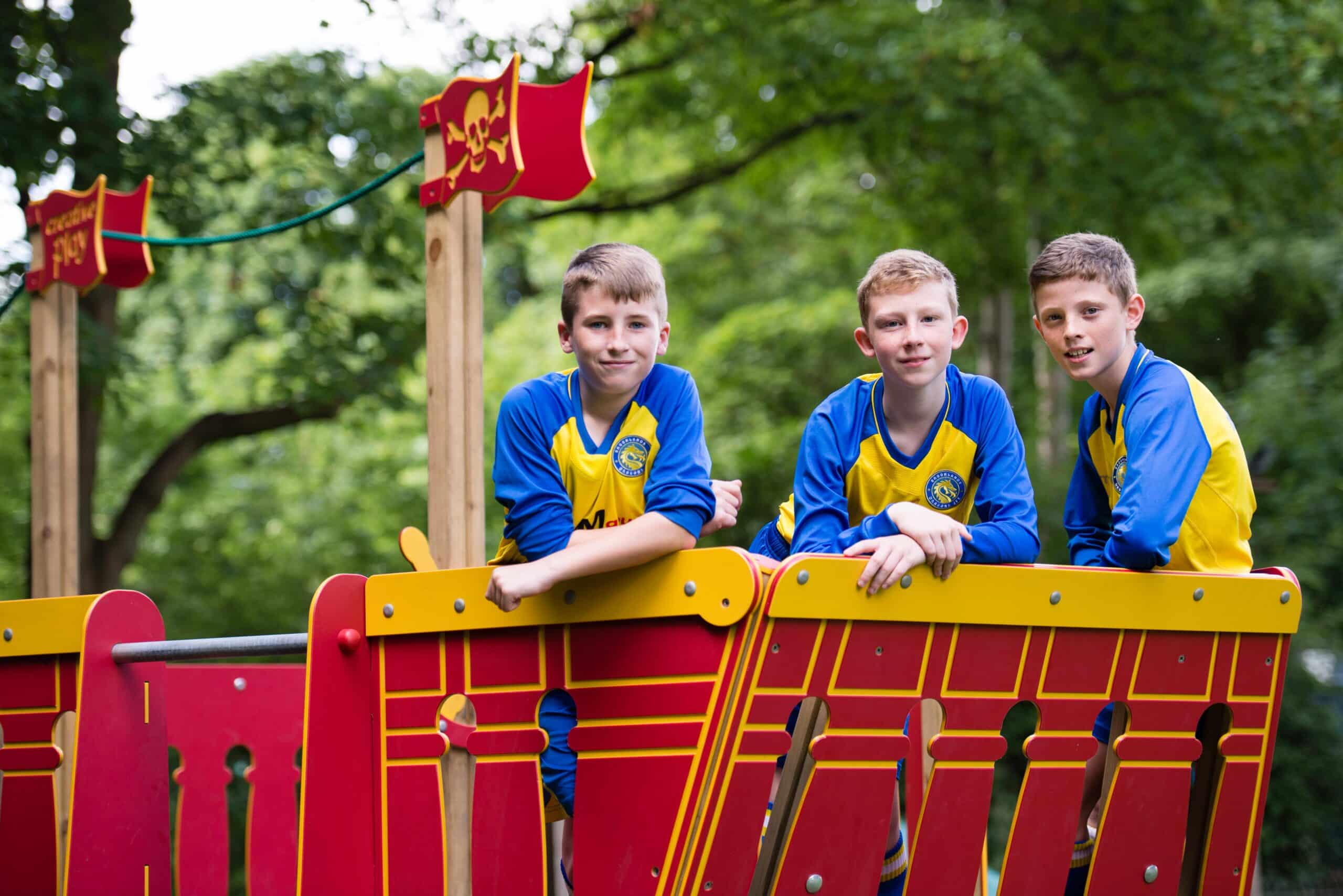 A wooden red ship in a playground with three boys in blue and yellow kits stood on top posing for the camera