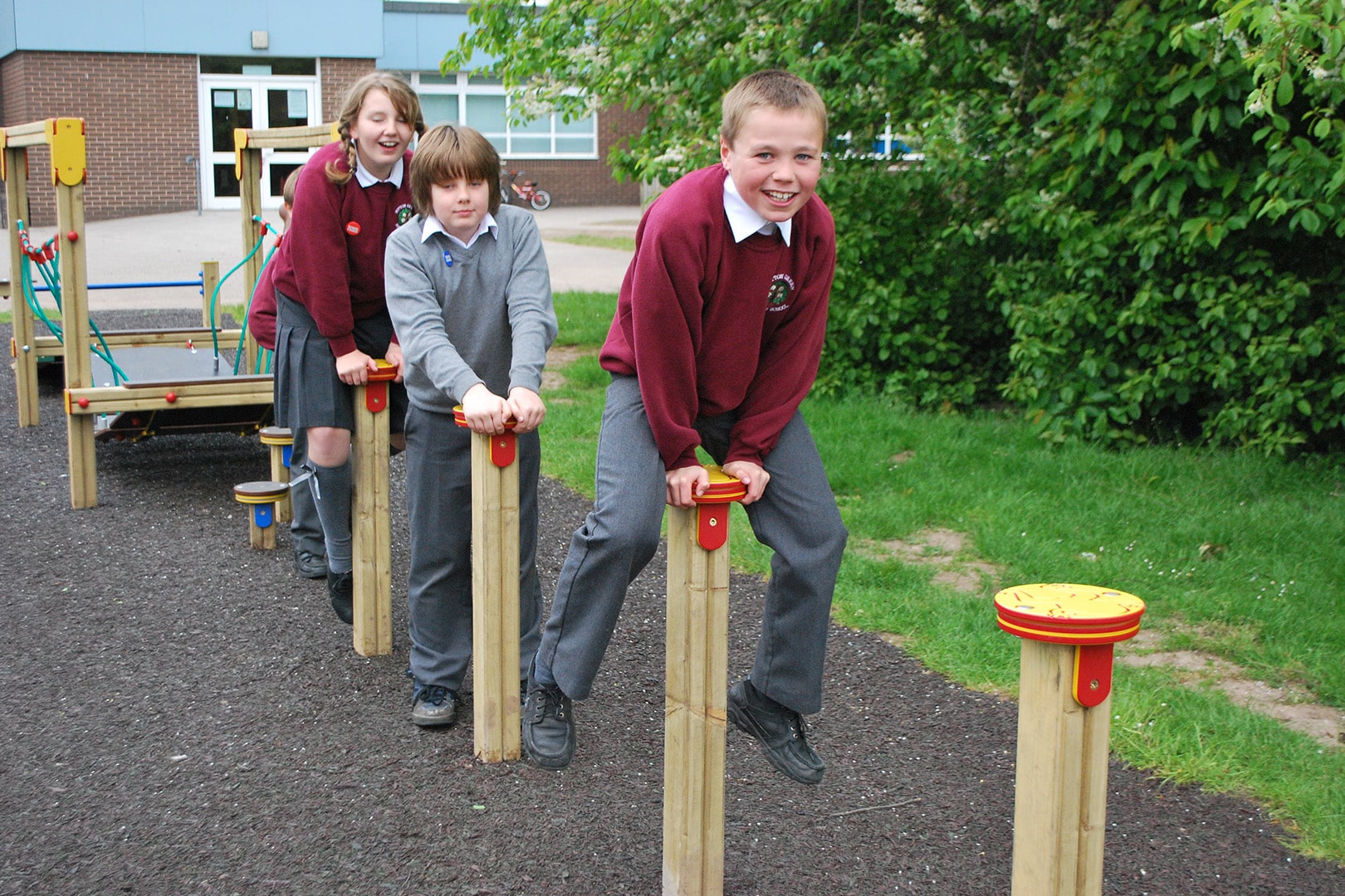 Three children, all in school uniforms, play leapfrog over wooden posts
