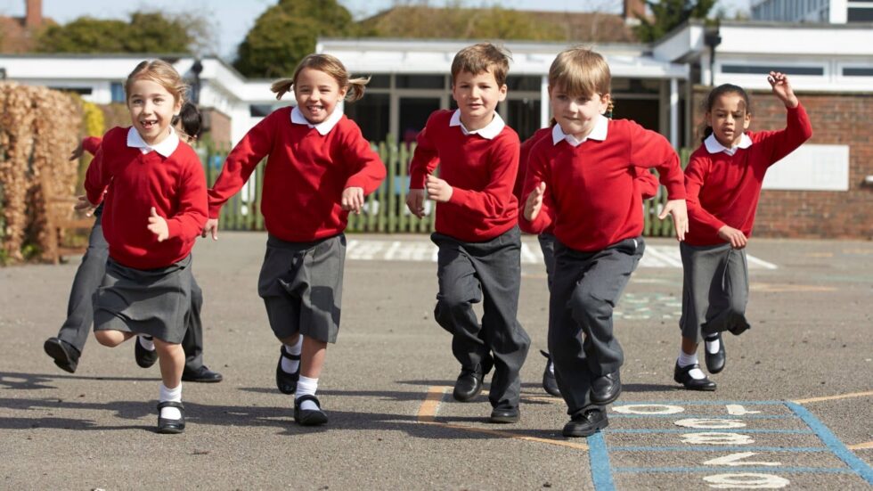 Children in a school yard wearing red uniform jumpers and grey trousers running towards the camera