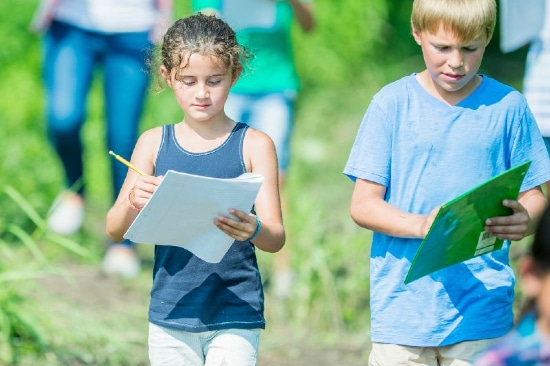 Children walking in a field on a sunny day holding papers and pens, making notes