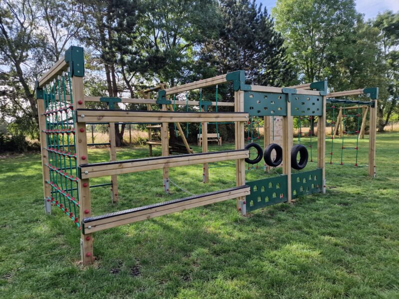 Wooden climbing frame in a park, includes green plastic panels and swinging tyres