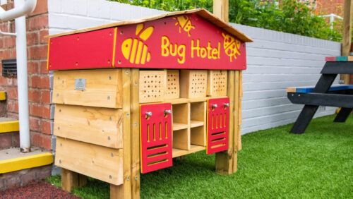 Wooden bug hotel with a red roof, sat against a play area wall outdoors