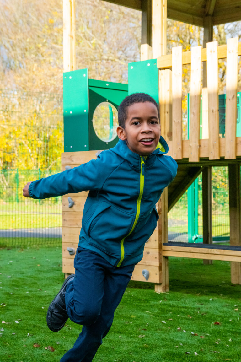 A young boy in a raincoat runs around on the green in front of a playtower