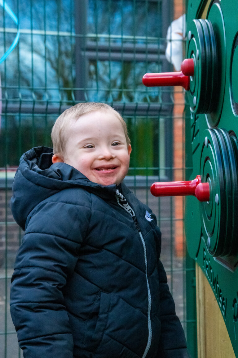 A boy with Down syndrome in a puffy navy coat looks to the camera with glee as he is about to embark on phone with an outdoor play panel with red handles