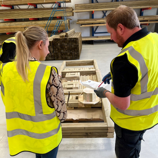 With their backs to the camera, a man and woman, both wearing high-viz vests consult a piece of paper over a stack of timbers in a warehouse