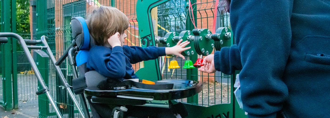 A child in a wheelchair plays with multi-coloured bells attached to an outdoor play panel