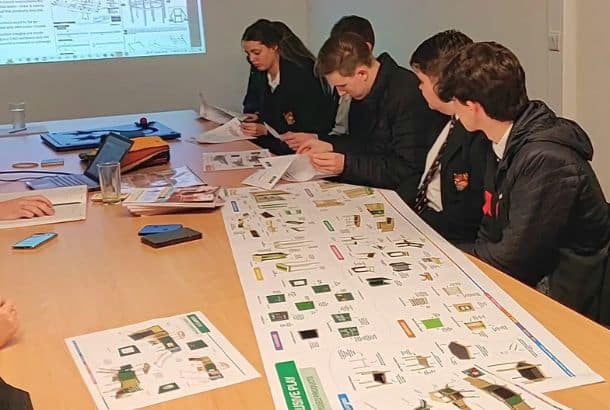 Pupils in uniform sit at a desk inspecting sheets of paper featuring product designs