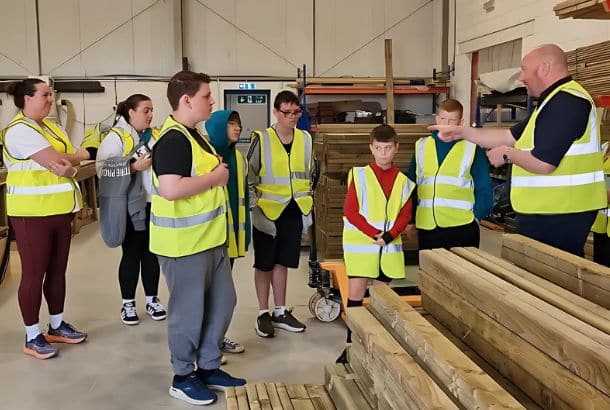 Several children in high-vis vests listen to a man talking surrounded by timber in a factory