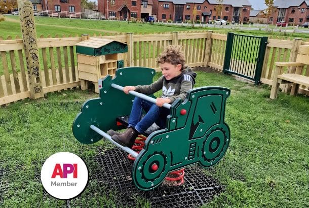 Boy on green tractor springer on grass with timber fence and housing in background plus sticker on the image which says API Member