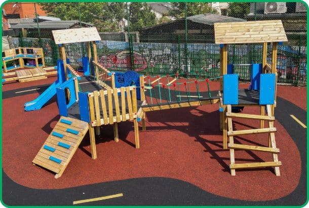 Timber play tower featuring two towers plus a hexagonal middle platform with blue panels sat on red surfacing in a school playground