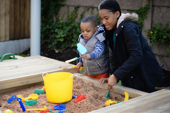 New Website Imagery - EYFS (1) This image shows a black mother and her young boy playing with a bucket in a sand box