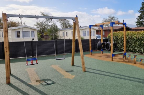 Swings at holiday park timber play towers and other play equipment on blue and red wet pour surfacing with caravans in the background