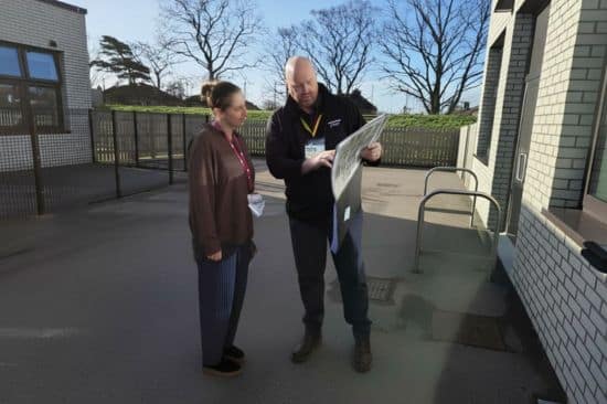 Man and woman stood in a grey school playground as he mansplains a design to her with sheets