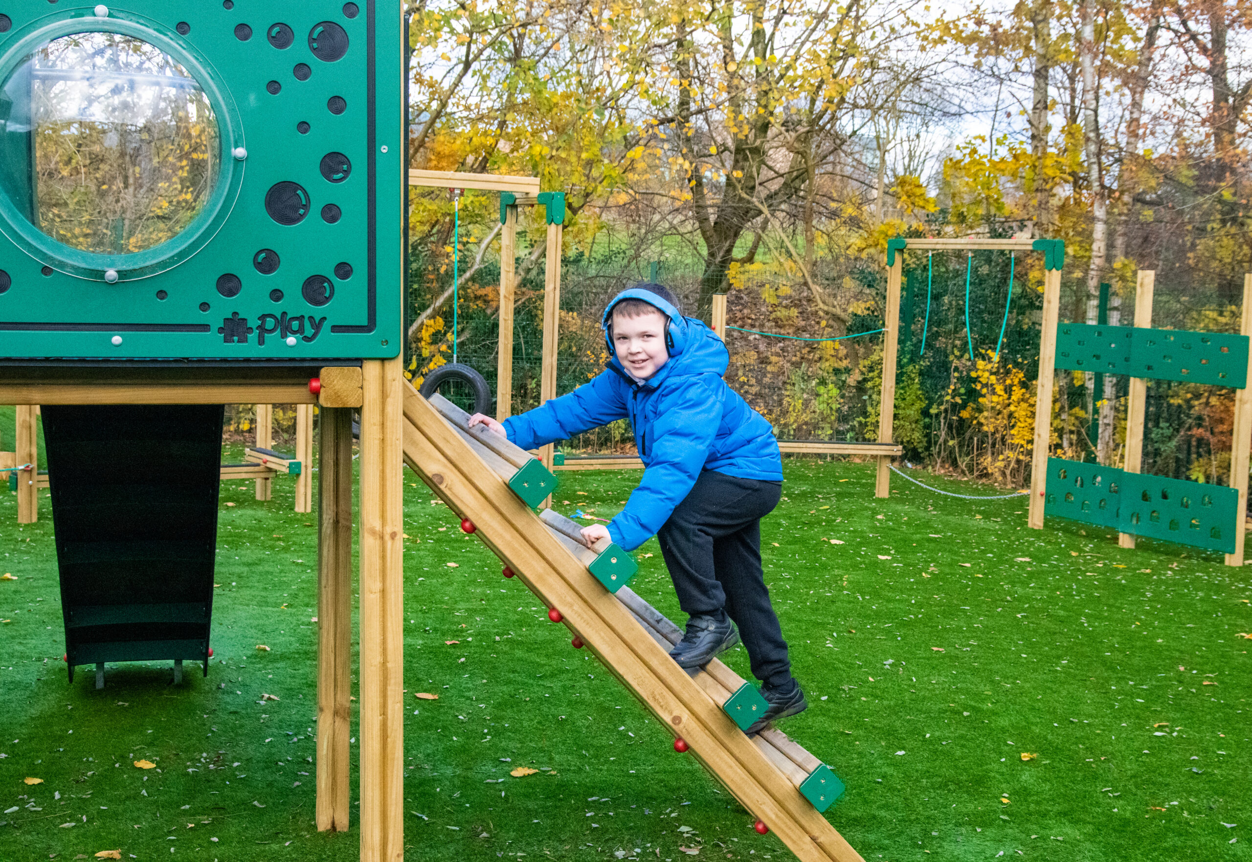 Boy in a blue jacket looks at the camera while he ascends a climbing platform to a wooden playtower. He is wearing blue headphones due to sensory requirements