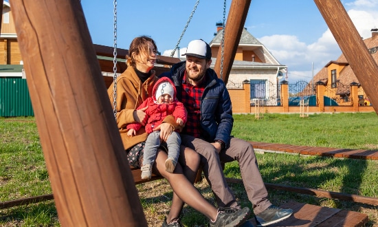 A mother and father holding a toddler as they all sit on a swing in the park