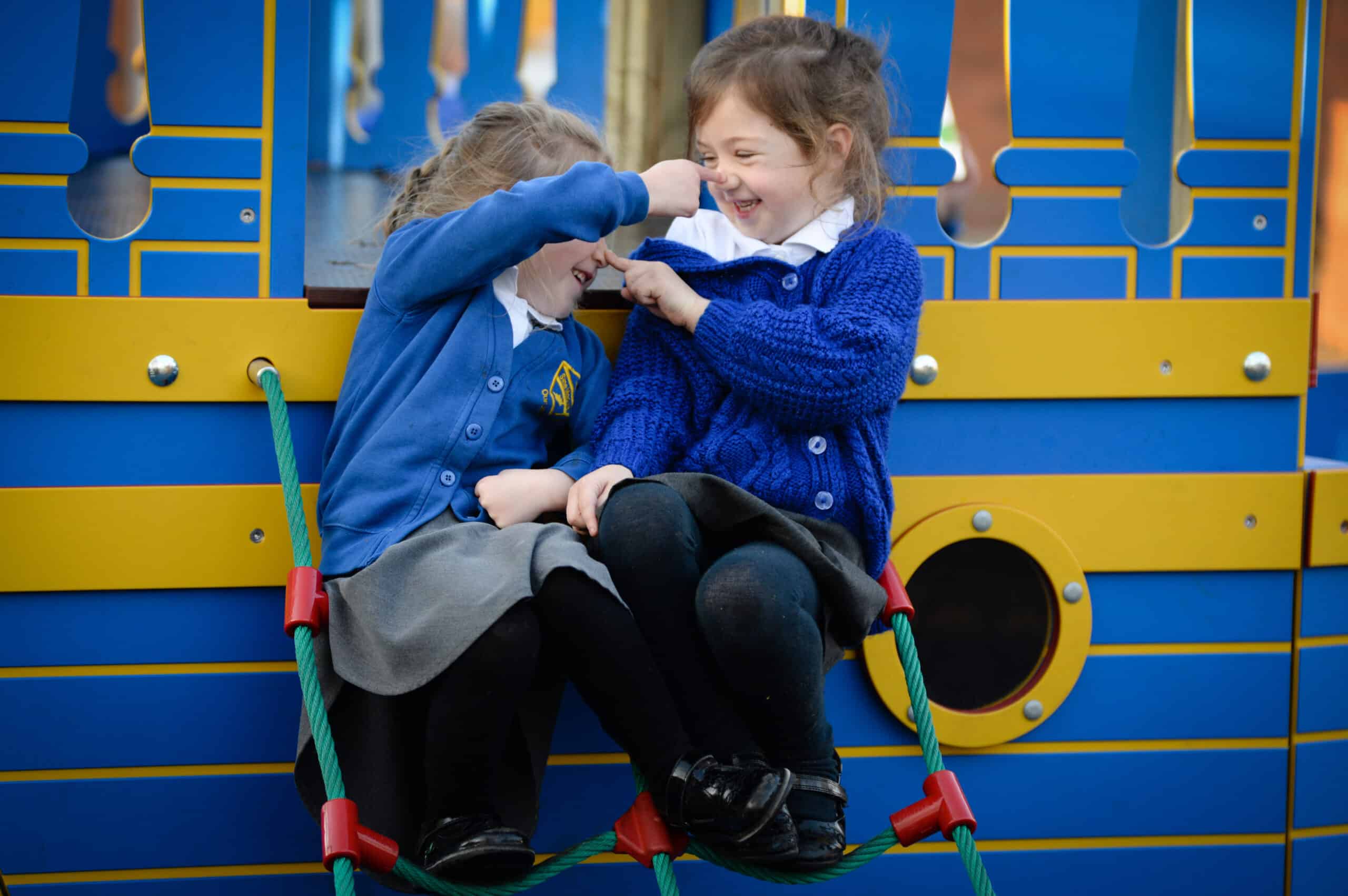 A blue and yellow boat made of wood in a playground with girl sat on it booping each others' noses