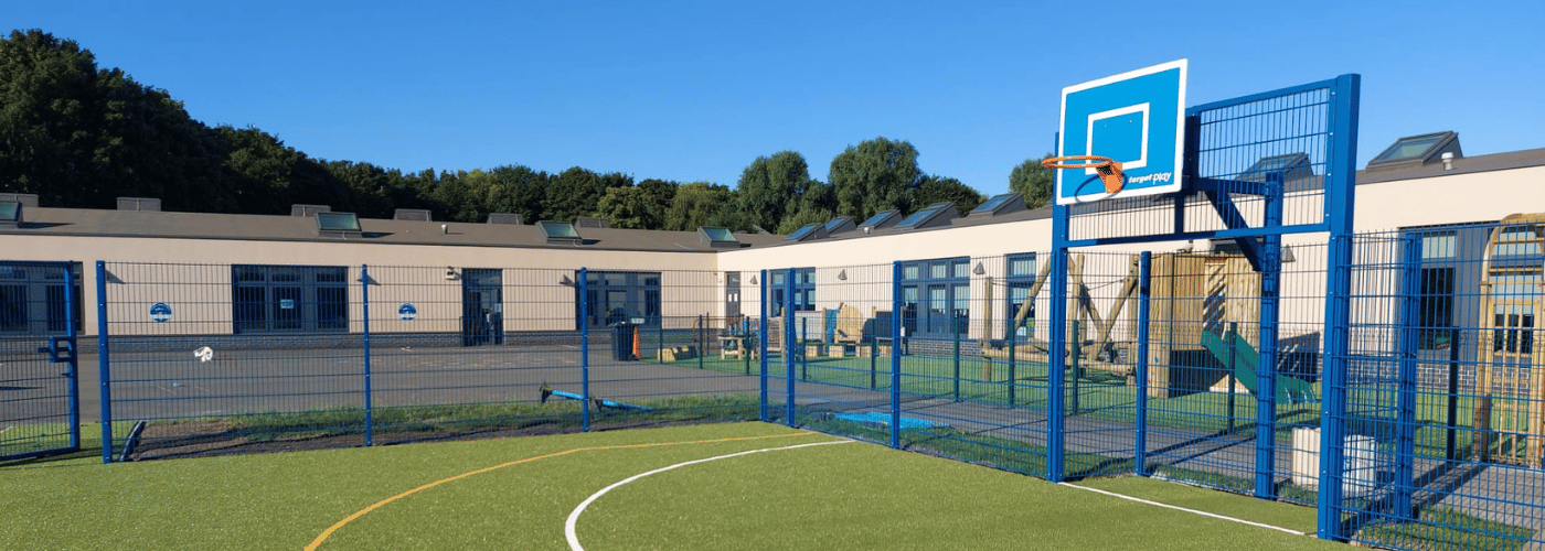 Blue fencing MUGA neatly fitting the corner of a school building, which can be seen in the background