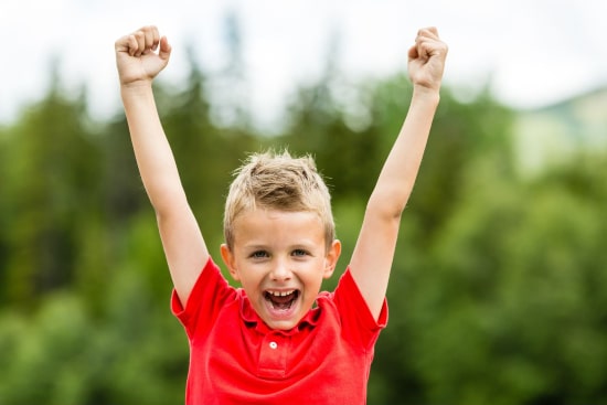 Little blonde boy wearing red raising his arms up in the air while smiling