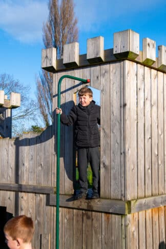 A young boy stand atop a wooden castle at a playground, contemplating sliding down a pole