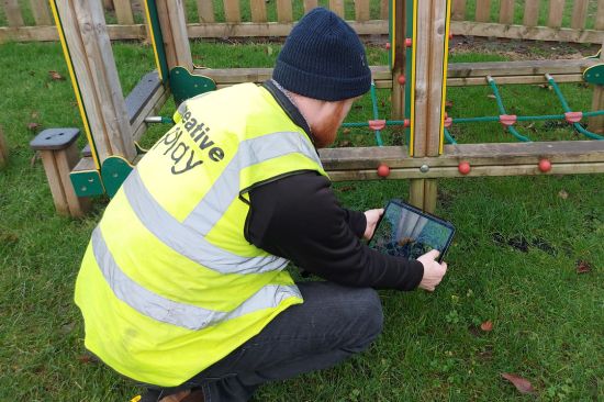 This image shows a male playground inspector, taking photographs on a tablet of repairs that need doing to playground equipment 